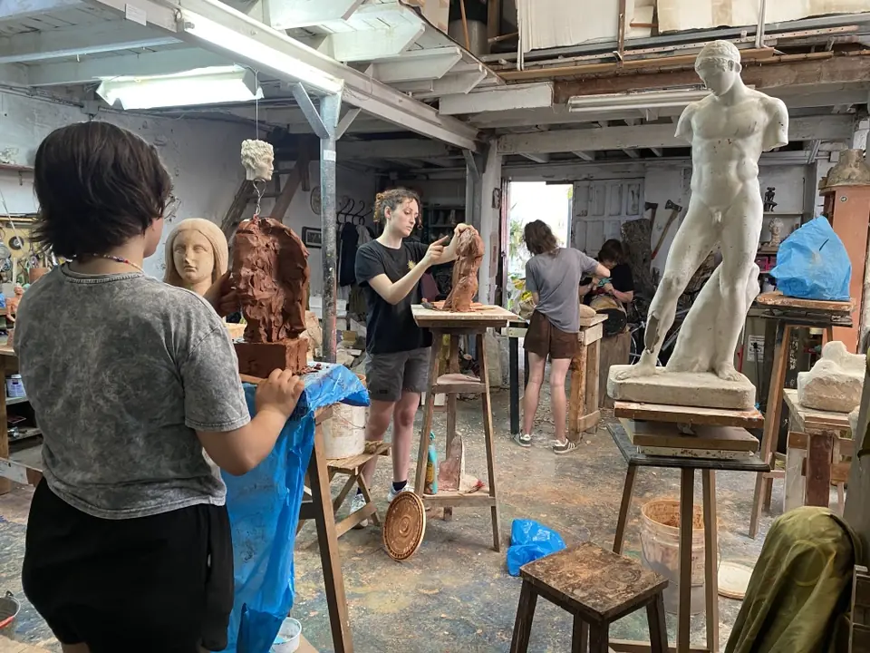 Alumnos modelando figuras en barro alrededor de una escultura clásica en el taller