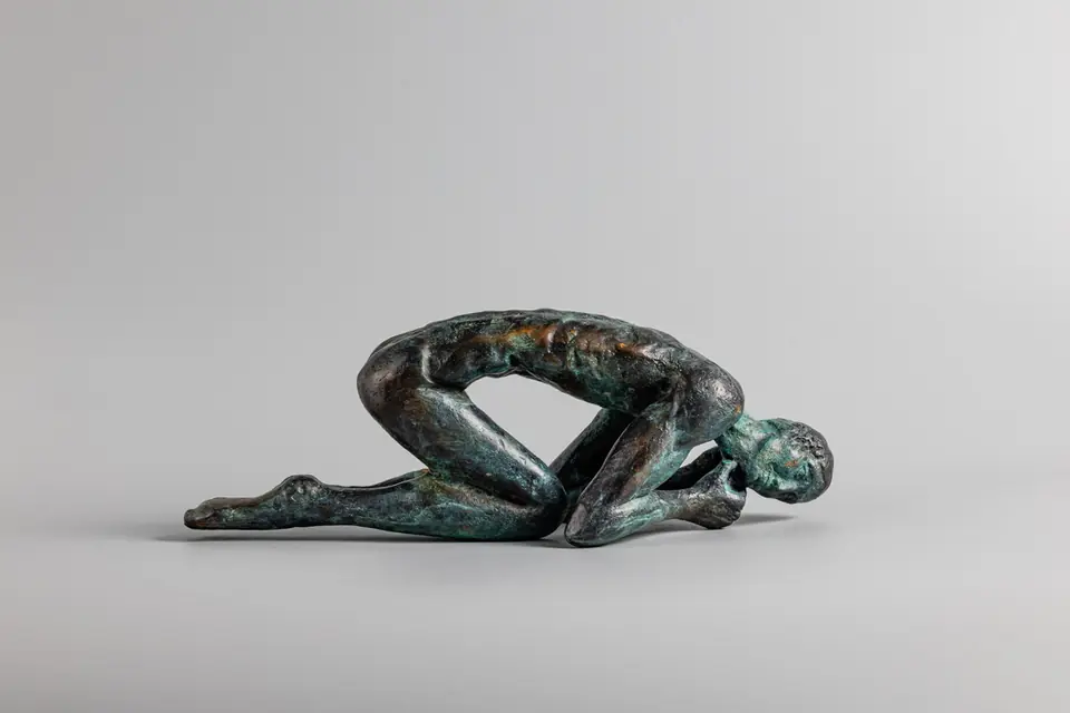 Figura humana encogida fundida en bronce con pátina verdosa sobre fondo gris claro